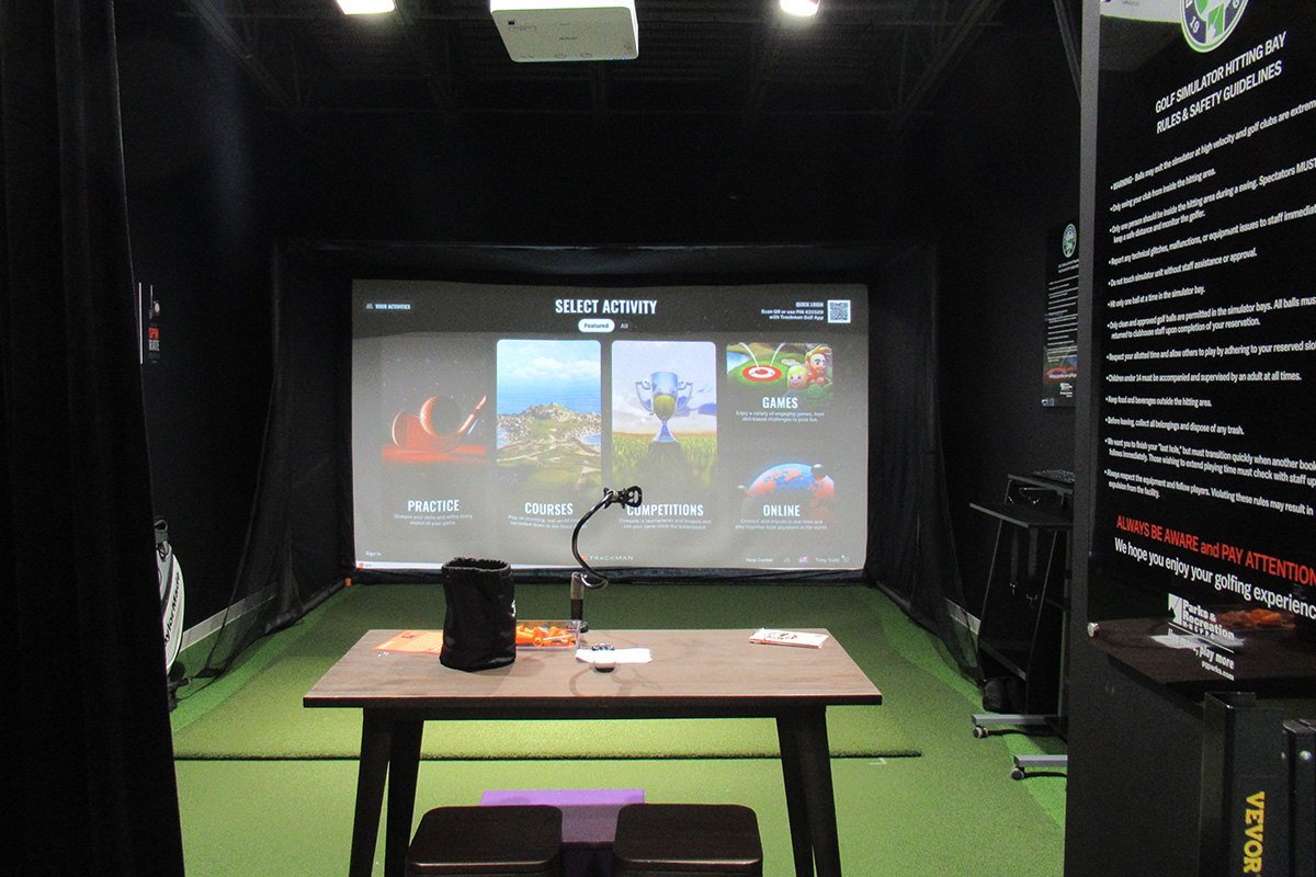A golf simulator room with dark walls, a large projector screen, and green turf on the floor. A large sign lists Golf Simulator Rules & Safety Guidelines.