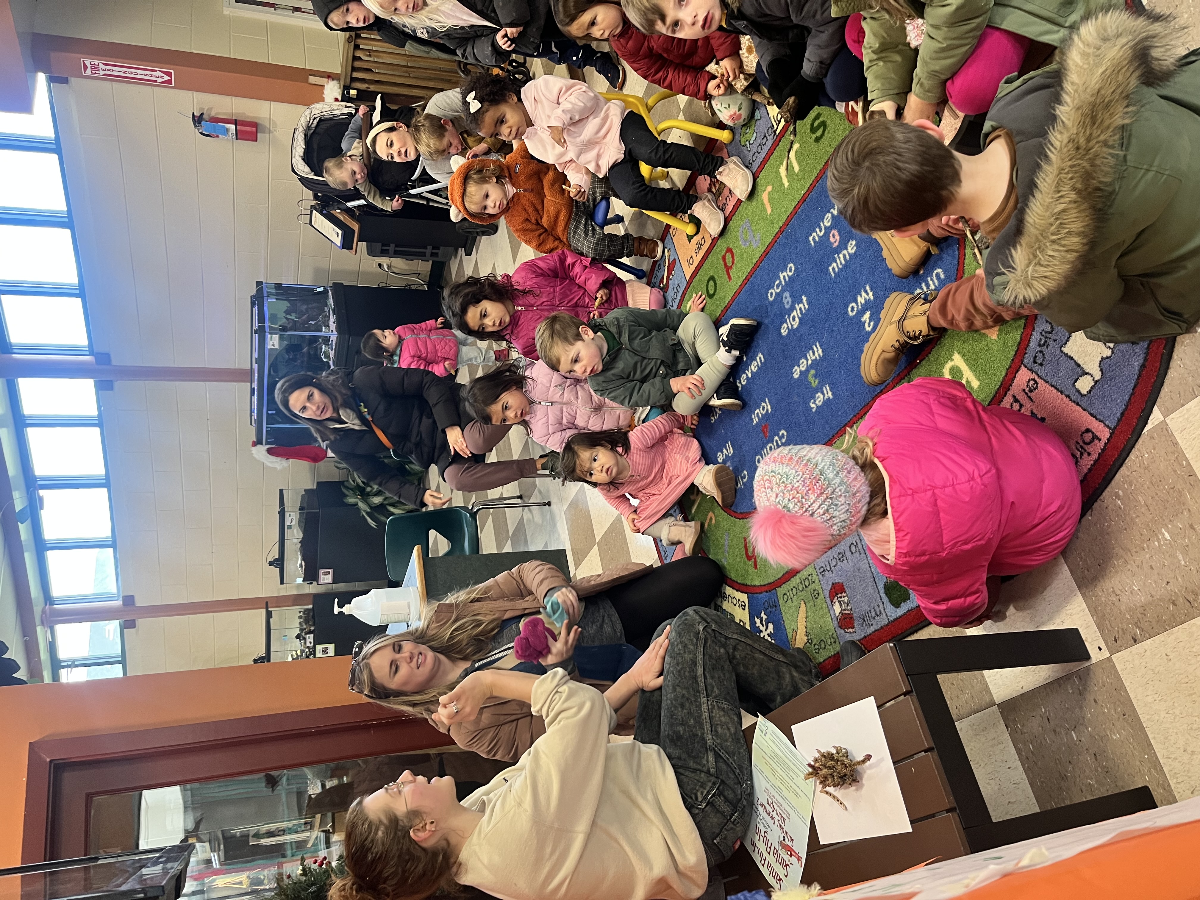 A person speaks to toddlers sitting on a colorful rug. Behind them are glass aquariums and terrariums along a curved wall with a row of windows at the top.