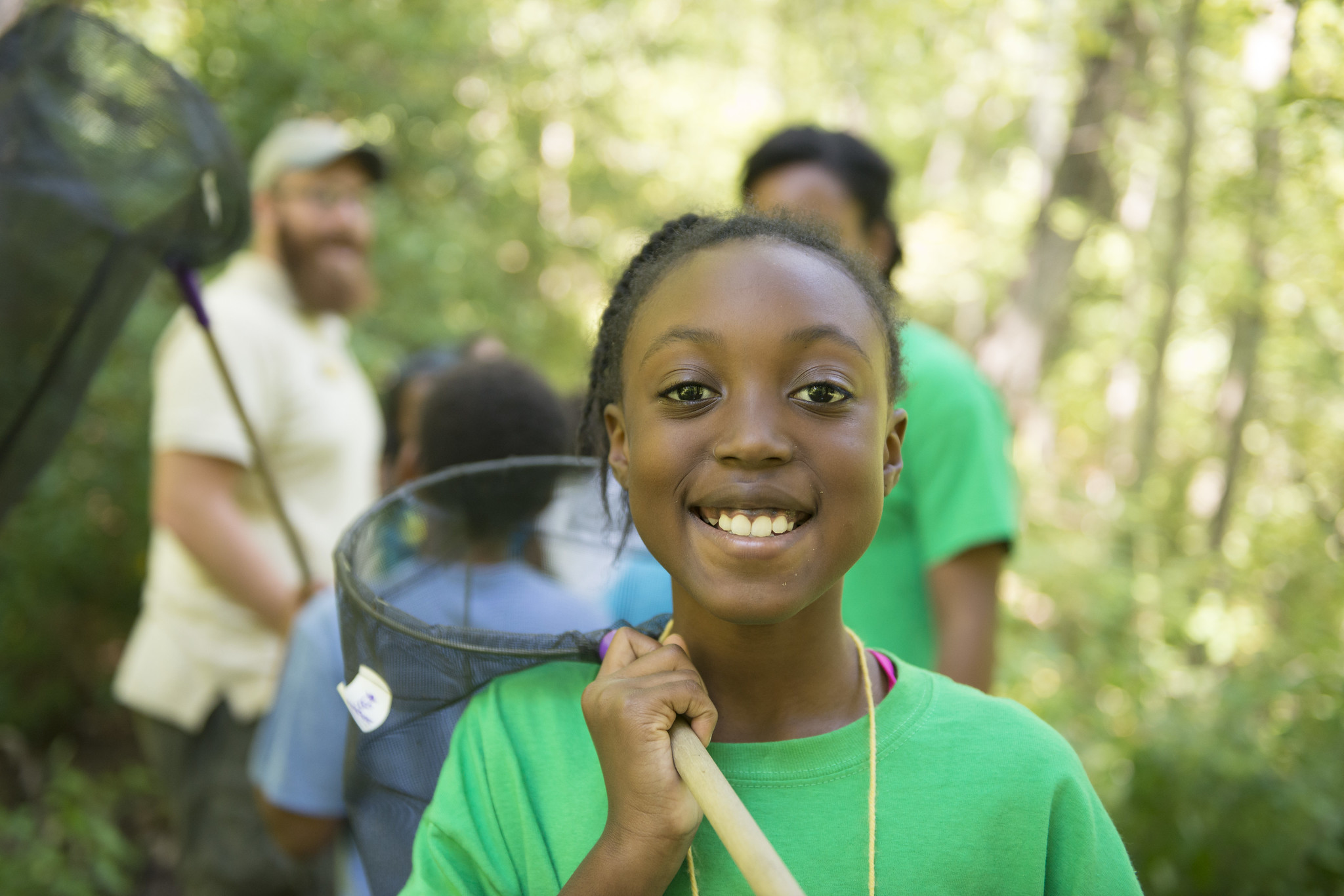 Close-up of a smiling girl in a green shirt holding a net on a long pole over her shoulder. Behind her is a wooded area.
