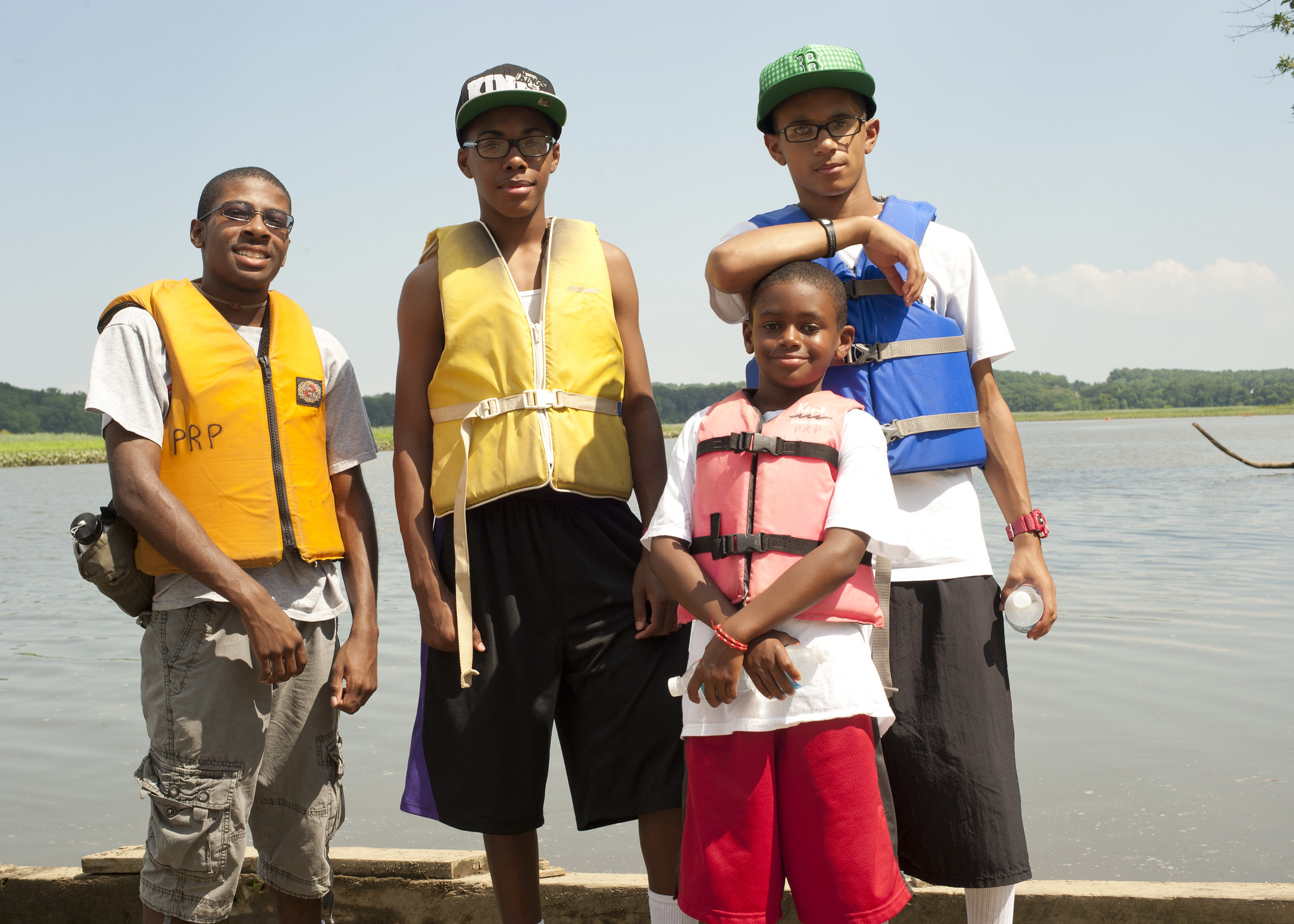 Four youths pose smiling in life vests on a dock next to a large body of water.