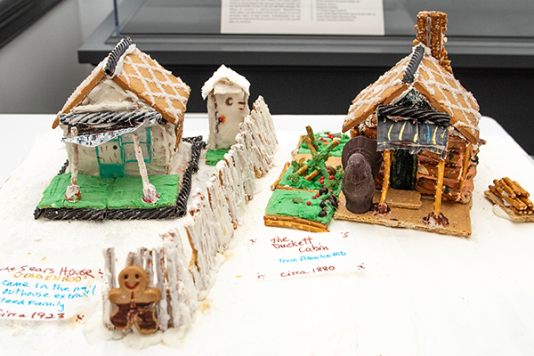 Two gingerbread houses with pretzel supported entryways. Both are adorned with black licorice decorations. There is a frosted pretzel stick fence between the two.