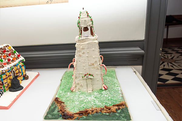 A tall white gingerbread lighthouse. It has candy cane supports and a chocolate bell and light at the top.
