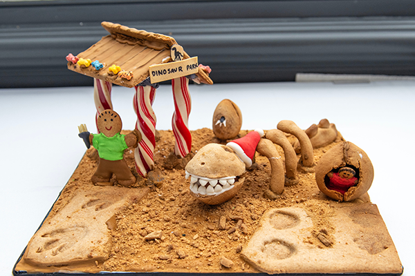 A gingerbread man under a candy cane stand with a sign that reads DINOSAUR PARK. He is standing in gingerbread crumb dirt and has found a giant festive gingerbread dinosaur fossil wearing a Santa hat.