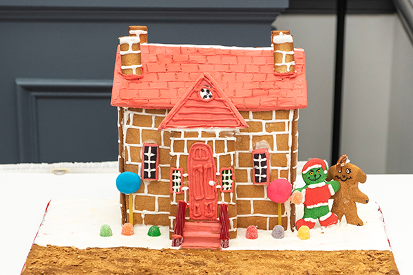 A gingerbread house with frosting lined brickwork, frosting roofing, licorice hand rails, and gumdrop shrubbery. The gingerbread Grinch and his gingerbread dog Max are standing to the side of the building.
