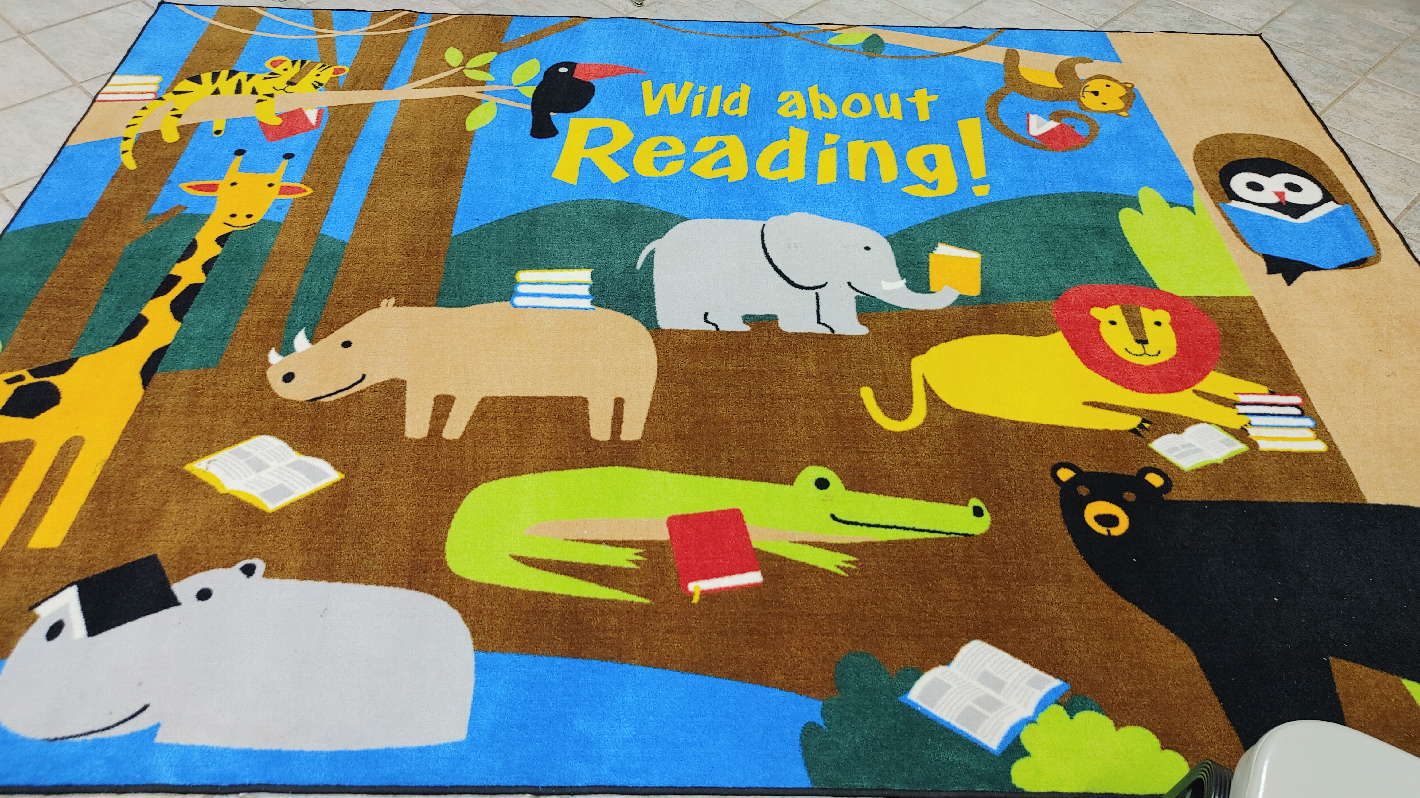 A colorful rug on a beige, tiled floor, with cartoonish images of wild animals in a nature setting with stacks of books, and the words WILD ABOUT READING at the top.