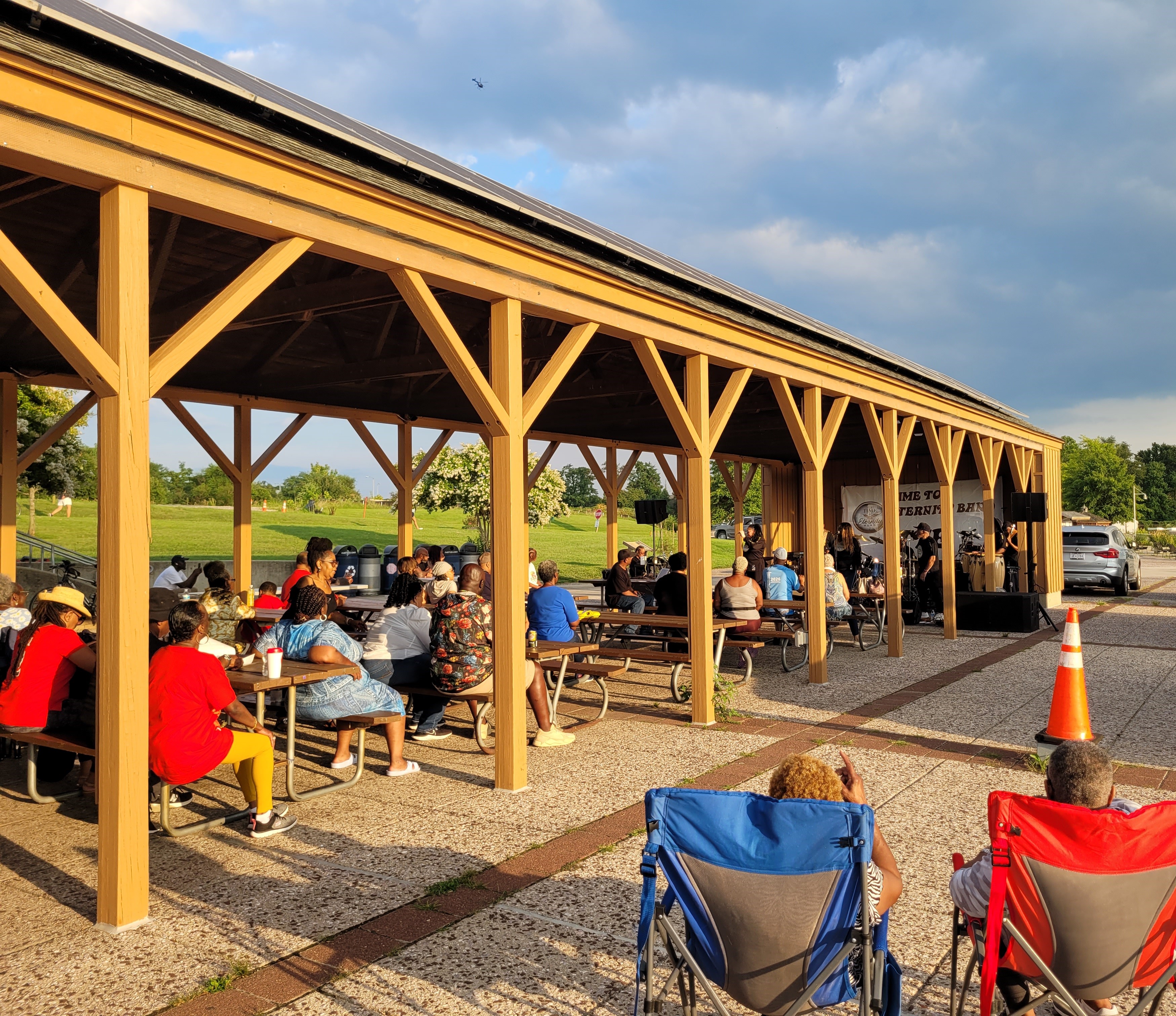 At sunset, spectators sit at picnic tables under a long, wooden, pavilion-style picnic shelter with a live band performing at the far end. Some people sit in lawn chairs on the concrete patio adjoining the pavilion.