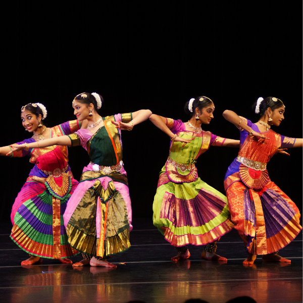 Four women wearing brightly colored dresses dancing on a dark reflective stage with a black background behind them