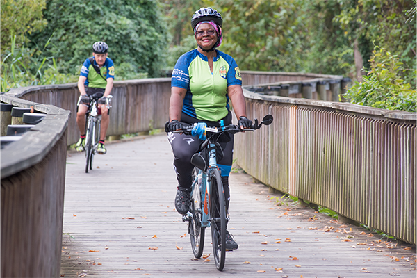 A woman riding her bike along a wooden bridge. She's wearing a helmet and a biking shirt. There is a man in the abckground wearing the same shirt also riding a bike.