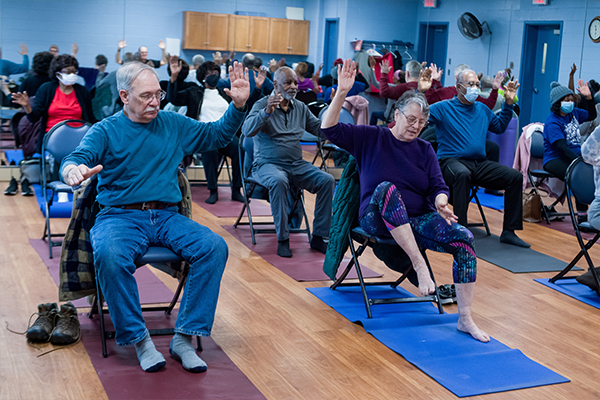 A group of seniors in a blue room with a wooden floor. They are all in chairs doing exercises in a class.
