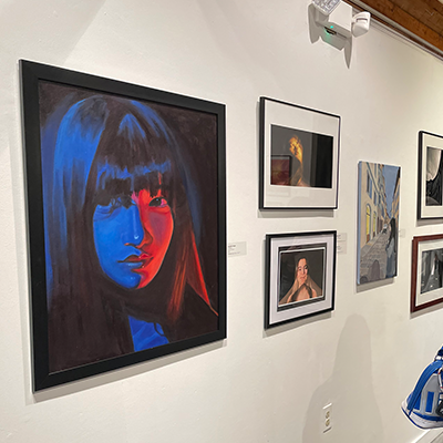 A white gallery wall with various artworks hanging on it. The most prominent is a large painting of a girl lit by very starkly contrasting red and blue lighting.