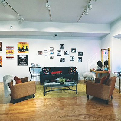 A couch, chairs, and tables are set up like a living room in a gallery corner with multiple framed images and large posters on the wall.
