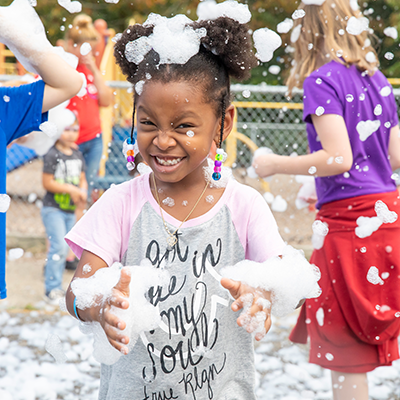 A girl grinning with soap bubbles covering her head and arms. There are other children playing in the bubbles behind her.