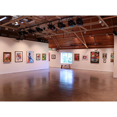 A room with white walls, a concrete floor, and a vaulted wooden ceiling. There is a window against the back wall casting a reflection on the shiny floor. There are various works of art hanging on the walls.