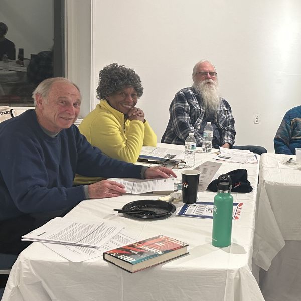Three seniors sit at a long table covered in a white tablecloth. In front of them on the table are print-outs, books, and beverages.