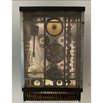 A wall-hung sculpture comprised of a cabinet-like box lit from within containing an assemblage of items made of glass, metal, and wood. One item is shaped like a hand.
