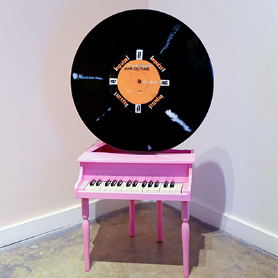 A small pink toy piano sitting in a corner with white walls and concrete flooring. There is a John Coltrane record positioned on top of the piano.