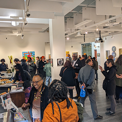 An art exhibition in a gallery with white walls, wood floors, and many people observing the works of art.