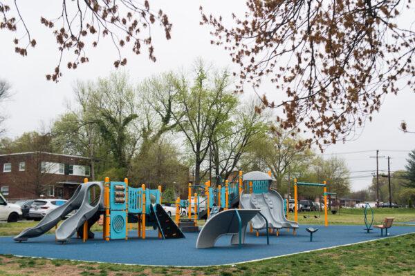 An outdoor playground with multiple play structures, featuring slides, platforms, stairs, climbing walls, musical instruments, games, spinning elements, and swings. Behind the playground are brick duplex houses.