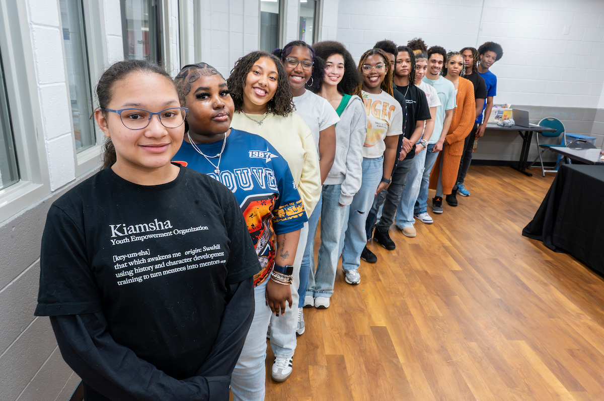 In a room with gray walls and a wooden floor, 14 smiling teens pose standing in a diagonal line facing the camera. The girl in front wears a shirt that reads KIAMSHA Youth Empowerment Organization.