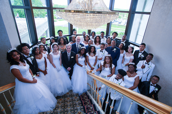 Several dozen people in formalwear pose on the landing of a large, carpeted stairwell with an ornate crystal chandelier above them, and a massive window behind them. Young women are all wearing white gowns and tiaras; young men are all wearing tuxedos. They all have corsages and boutonnieres with white flowers.
