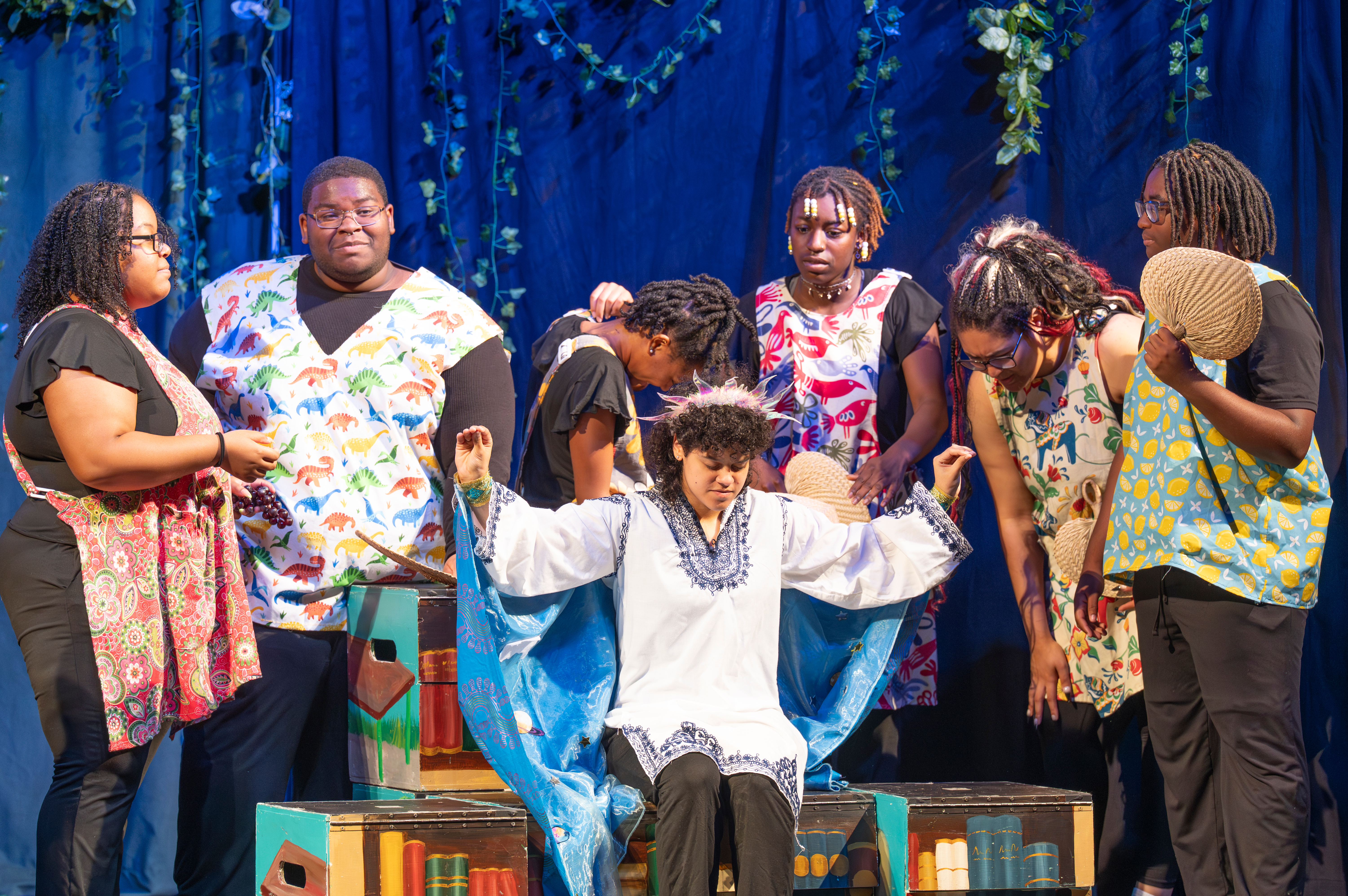 A group of teen actors performing on stage in patterned costumes around a seated cast member against a blue backdrop.