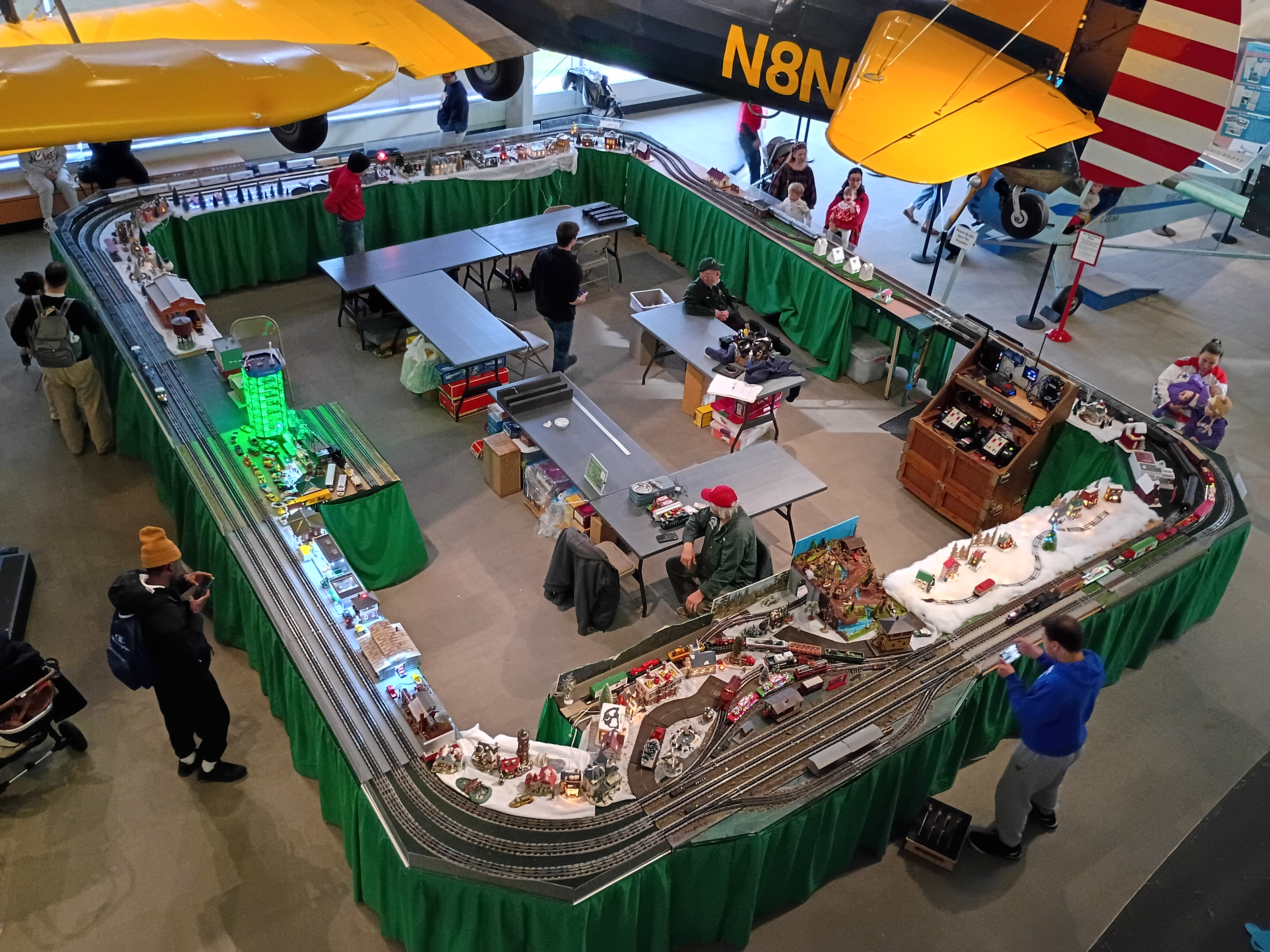Model train display inside an aviation museum with miniature buildings, tracks, and visitors beneath suspended vintage aircraft.