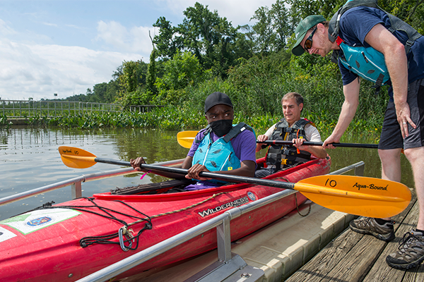 Two men sit in a red tandem kayak on a kayak/canoe launching pad attached to a wooden dock, about to enter a body of water. Both of them wear life vests and hold kayak paddles. A man on the dock is leaning down and holding onto the paddle of the man in the back.