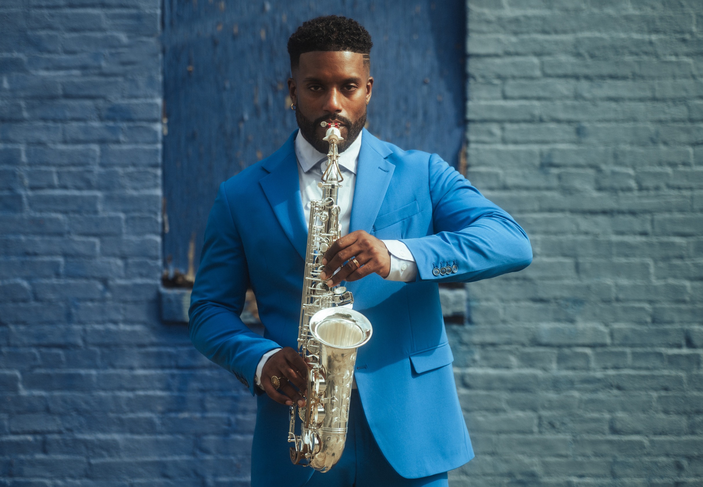 A man in a blue suit plays a silver alto saxophone in front of a brick wall.