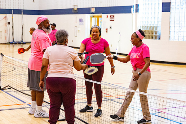 Four people standing in-between a net holding pickleball equipment. They are all wearing pink attire.