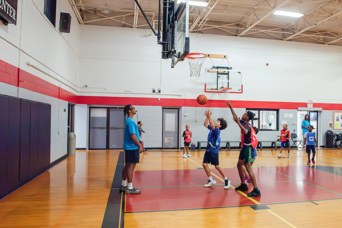 Boys in red and blue numbered smocks play basketball in a gymnasium while a staff member watches with a whistle in his mouth.