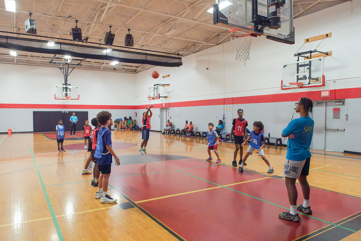Ten boys in red and blue numbered smocks play basketball in a gymnasium while a staff member watches with a whistle in his mouth.
