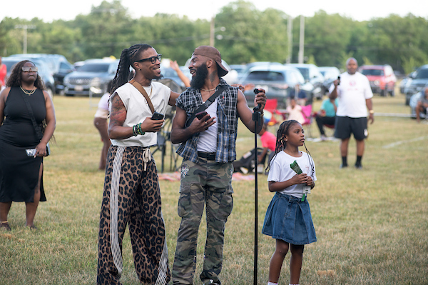 Two performers carrying microphone standing in a field beside a child during an outdoor festival.