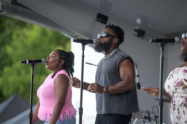 Three vocal performers sing into microphones on an outdoor stage during a live music performance.