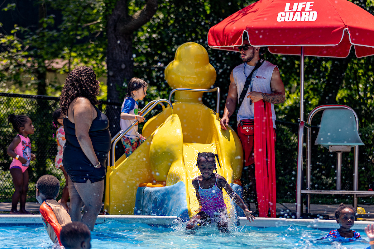 Small kids line up to go down a small, yellow, duck-shaped waterslide next to a lifeguard stand.