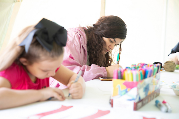 Under a white event tent, a girl and a woman sit coloring with markers.