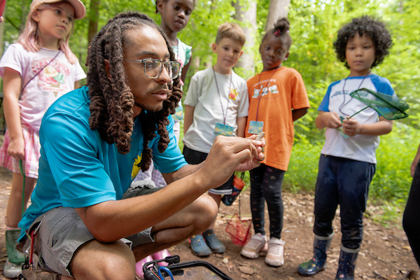 A camp counselor shows a small lizard to a group of curious children in a wooded area.