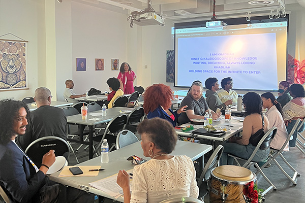 People sit at tables set up in an art gallery while a standing woman presents a slide presentation.
