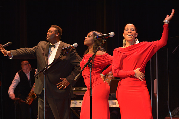 A man in a dark suit performs on a stage next to two women in red dresses.
