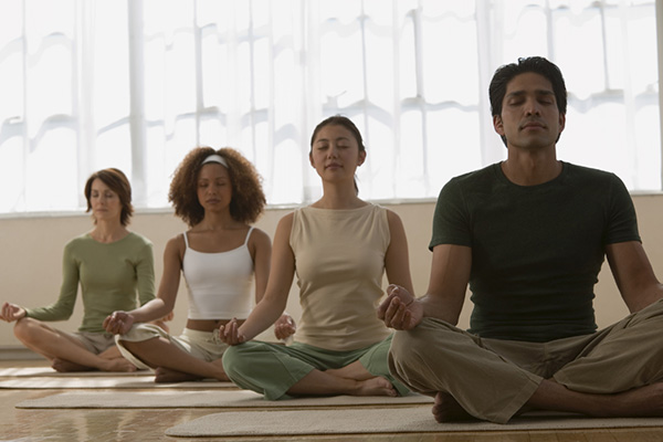 Four adults sit in the lotus pose with their eyes closed in a yoga studio.