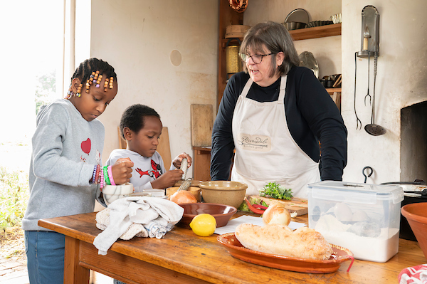 A staff member in an apron speaks to two kids who are trying out marble mortar and pestles in an 18th-century kitchen.