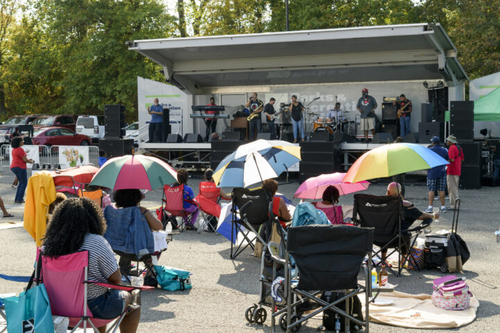 A band performs in a mobile bandwagon in a park, with audience members sitting in folding chairs on the pavement. Several use colorful umbrellas for shade.