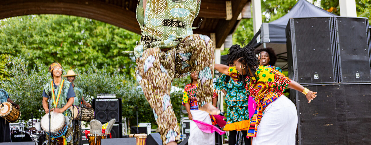 Performers in colorful patterned attire dance and play drums on an outdoor stage during a cultural festival performance.