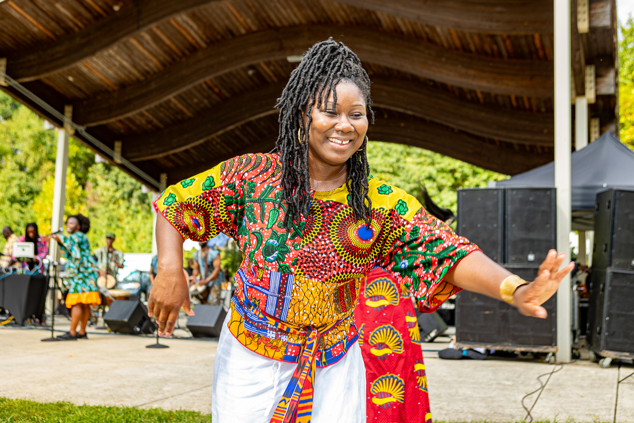 Performer in a brightly patterned outfit dances near an outdoor stage, with musicians, speakers, and festival attendees visible in the background.
