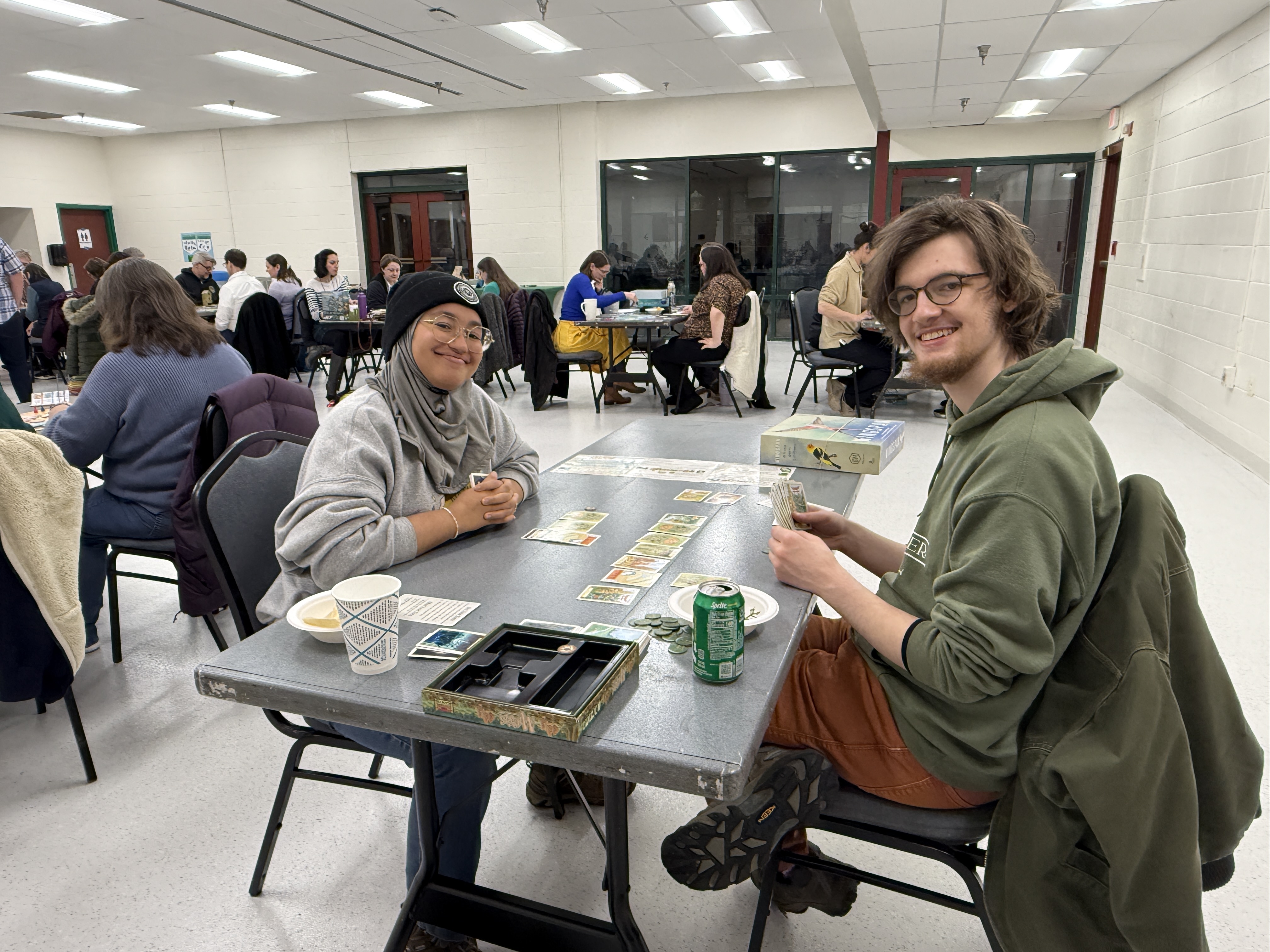 Two smiling adults sit at a table playing a card game. Behind them, the room is full of people sitting around tables playing board games.